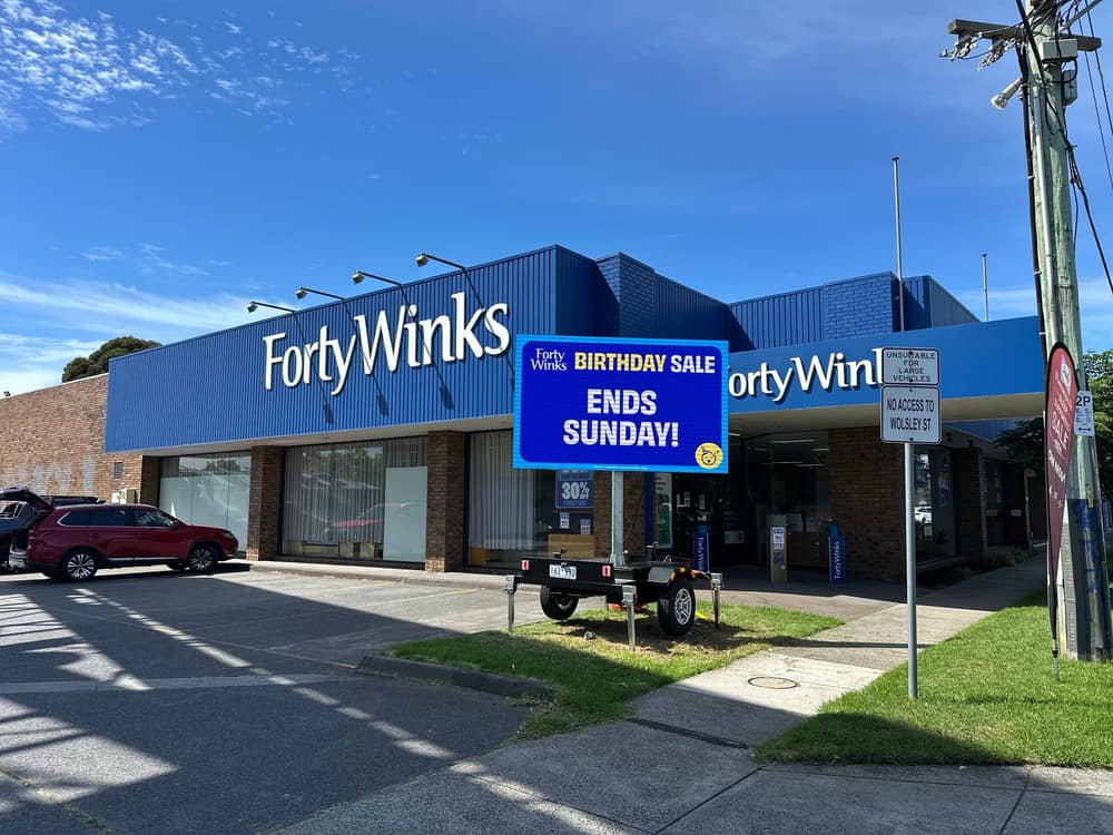 Ozzy's full-colour LED trailer sign parked kerbside outside a Forty Winks store in daylight, showing a blue 'Birthday Sale' promotion