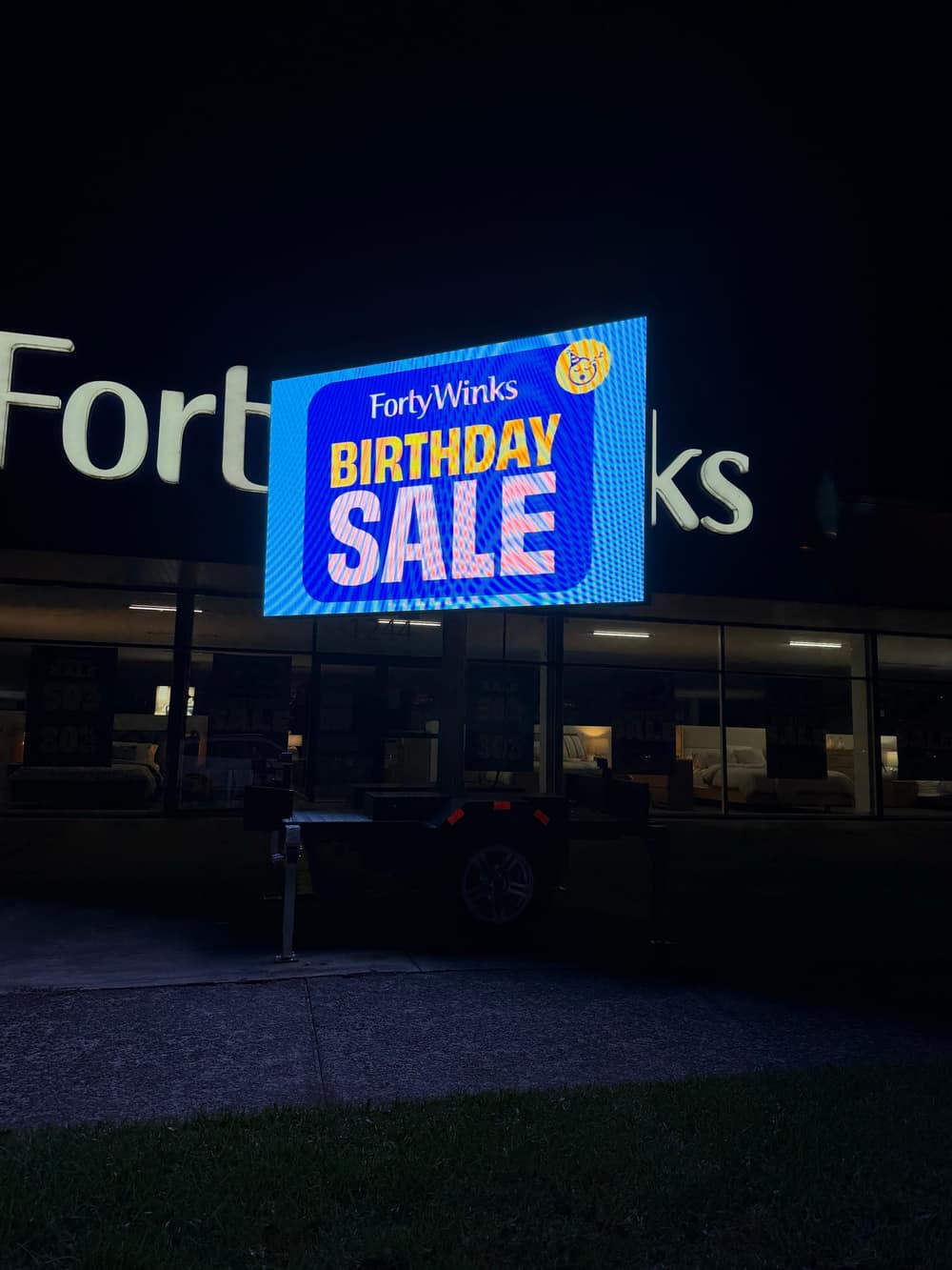 The same LED trailer sign lit up after dark outside the store, the 'Birthday Sale' artwork still clearly readable at night